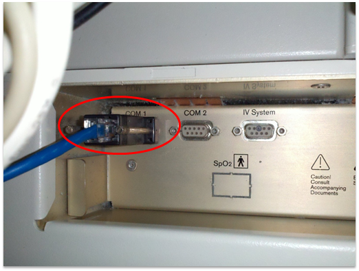Dräger anesthesia machine rear connector panel — COM 1 (circled in red) with a serial cable attached, beside COM 2 and IV System